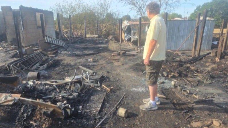 A man in a cream t-shirt and khaki shorts is standing in his garden that is destroyed by fire. Everything is black and burnt and there is rubble everywhere