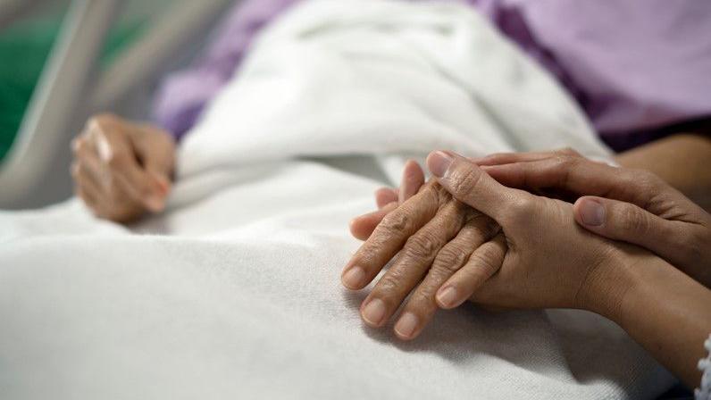 Someone holds a hospital patient's hand in their hands, with a blurred background of someone in lilac clothing in a hospital bed, under a white sheet