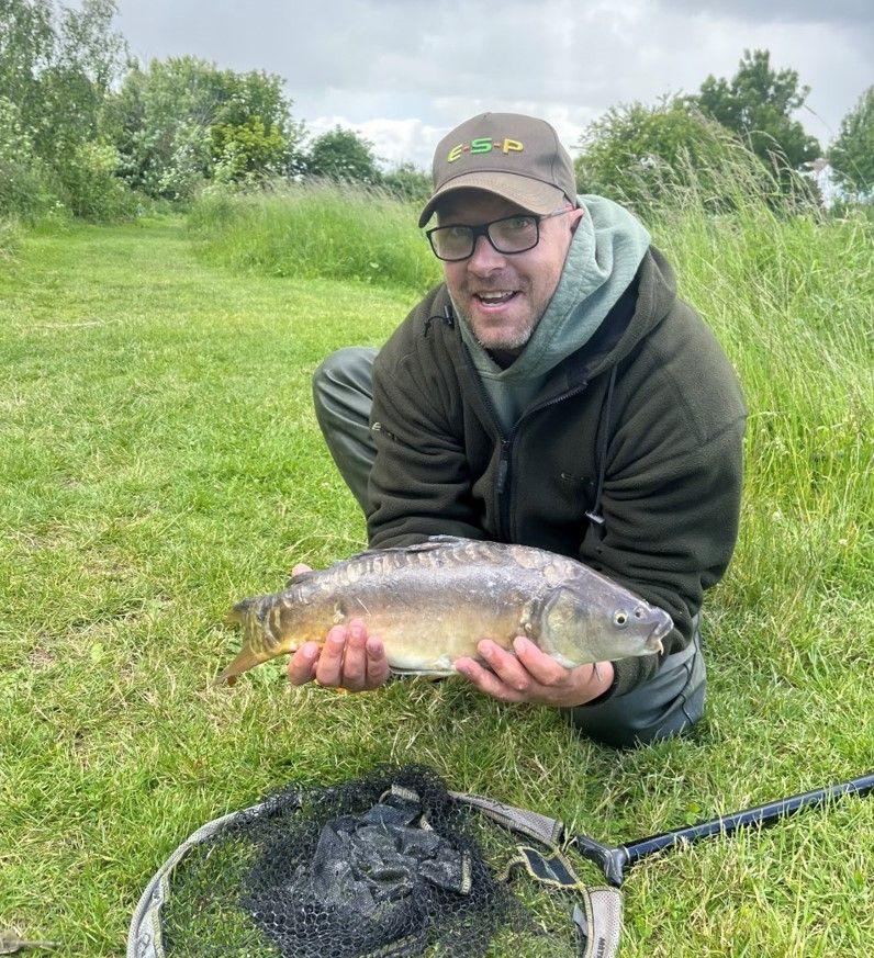 A man wearing a baseball gap, glasses and green clothing crouches next to a fishing net laid down on the grass. He is holding a fish with both hands as he looks to the camera