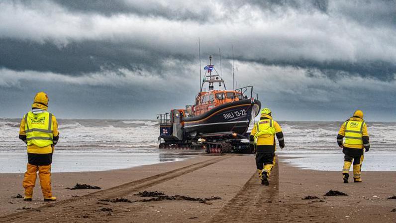 Three RNLI crew members in orange, black and yellow all-weather gear walk on a sandy beach towards an orange and black lifeboat as it is towed into the waves beneath a dark, stormy sky.
