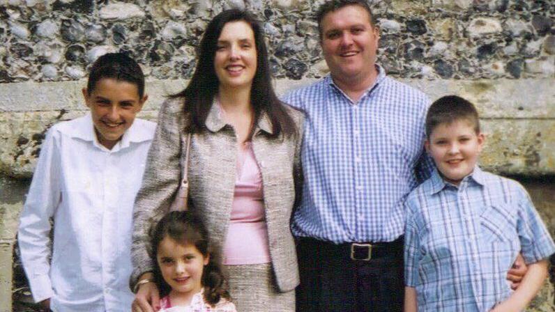 A family photo taken against a flint wall. Oliver had short dark hair and wore a white shirt. He is standing next to his mother, who had long dark hair and is wearinf suit with a pale pink top and pink handbag on her shoulder. She has her hand on her young daughter's shoulder, who also has dark long hair, pulled back into a ponytail. Thomasina's late husband had his harm round her and had short, fair-ish hair and was wearing a blue and white checked shirt. He had his other arm around his son Elliott, who is also wearing a blue checked shirt. The family are all smiling at the camera.