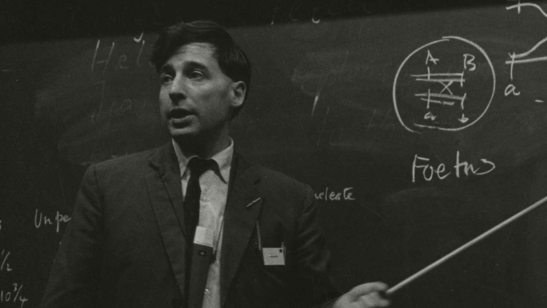 Prof Edwards at a symposium in Berlin on sperm capacitation. He is formally dressed and stands beside a blackboard filled with diagrams and handwritten notes. He gestures with a long pointer toward a circular schematic and a graph labeled with variables 'a' and 'b'. The board also includes references to developmental stages, such as 'Foetus' and numerical ranges.