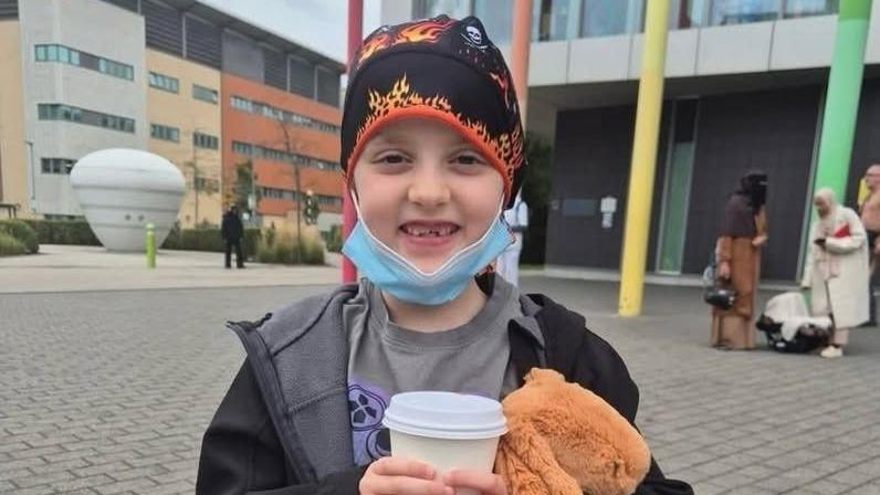 A little girl stands in front of a building. She wears a black and orange bandana on her head and has a face mask pulled down to show her smiling. She holds a takeaway cup and a cuddly toy.