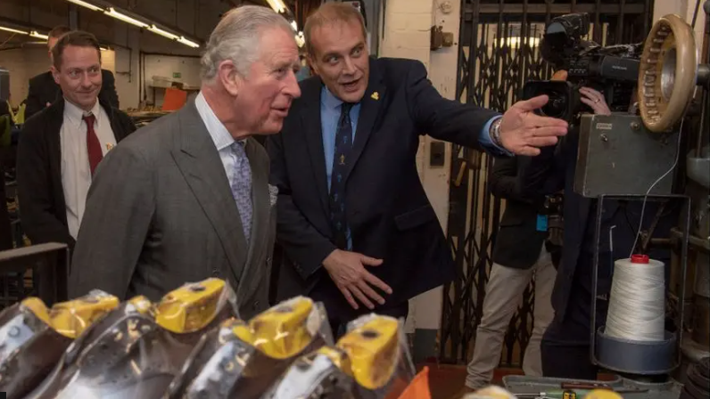 Inside a shoe factory King Charles is wearing a brown suit with a lilac-coloured tie. He is standing next to another man in a navy suit who is pointing to a manufacturing machine. Several shoes in the process of being made are on display