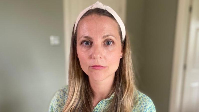 Julie Imrie looking straight ahead with a neutral expression. She has long, dark blonde hair and is wearing a white headband and a green shirt with blue on.