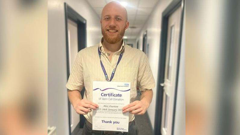 Alex Overton is smiling as he stands in a corridor holding his certificate. It says 'certificate of stem cell donation' and includes the words 'thank you'.