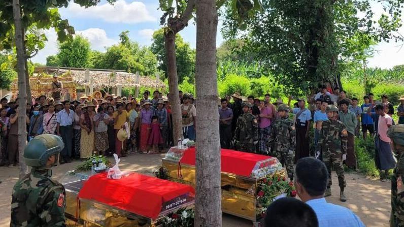 Villagers gathered around two coffins, with red cloths draped on top of them