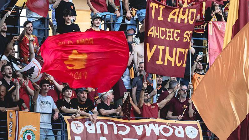 Roma fans in red club shirts, holding up banners and flags in a stadium