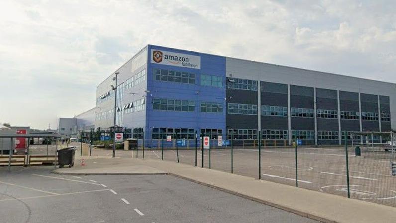 A large warehouse building, viewed from a car park. The corner of the building has blue cladding and an Amazon company logo on a white background at the top.