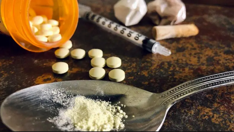 A close up of pills spilling from a bottle, a spoon with a powder on it, a syringe and the butt of a cigarette or joint.