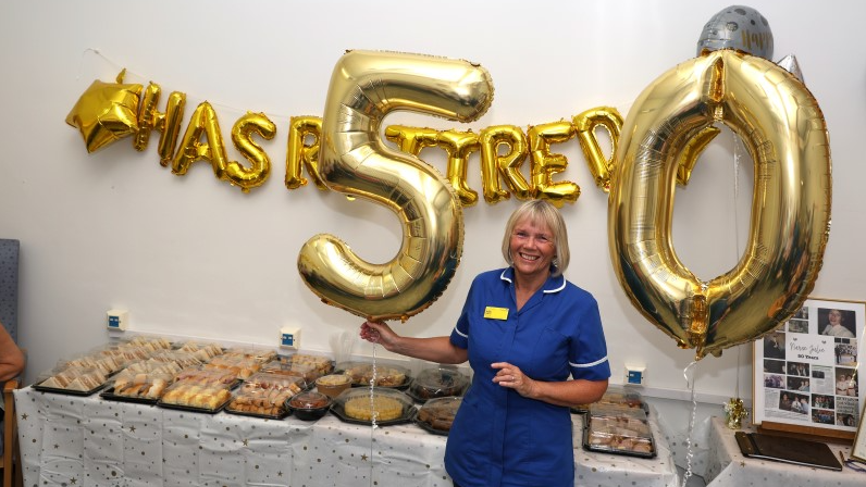 Julie is wearing her uniform and is stood in front of balloon letters spelling out that she has retired as she holds the numbers 5 and 0 in front of cakes and various treats.