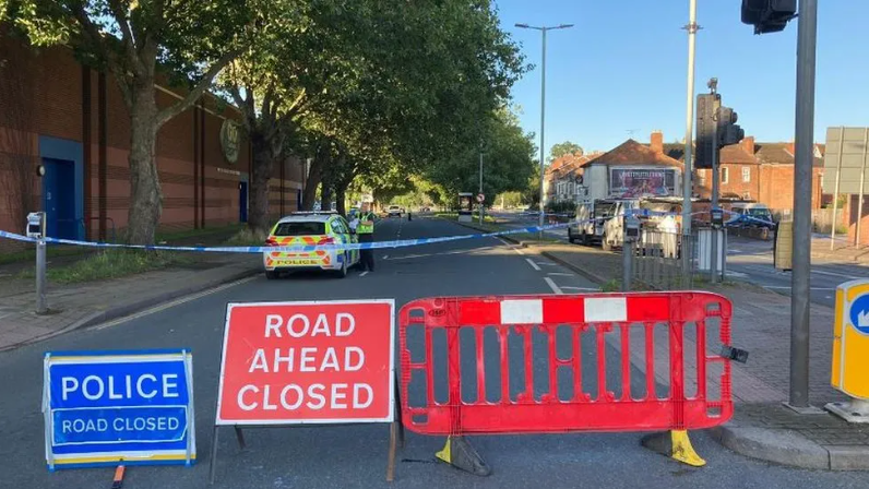 A blocked off road with police tape, a barrier, a red Road Ahead Closed sign and a blue Police Road Closed sign. There is a police car and officer in the background