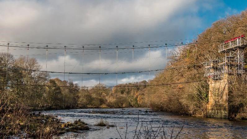 Whorlton Bridge. The components have been removed from the bridge. A network of cables stretches over a river in its place. To the right, the bridge's remaining stonework can be seen on the river bank. There is scaffolding on top of it.