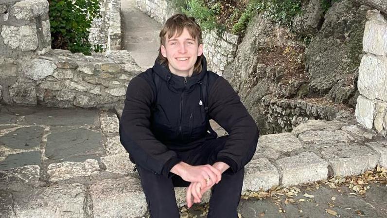 A young man with blonde hair smiles as he sits on a stone ledge at a ruined site. His hands are resting on his knees and he is wearing a black coat and black trousers