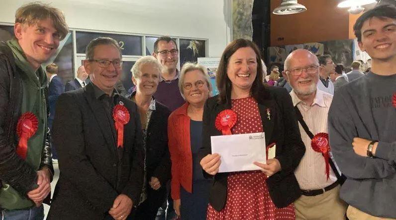 Julia Buckley is wearing a red dress with white polka dots and a black jacket. She is joined by a group of Labour Party staff and they are all wearing red rosettes, as she celebrates her general election win.