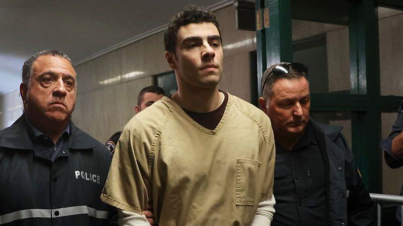 Luigi Mangione wearing a pale yellow prison jumpsuit being escorted by two prison officers on either side of him