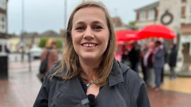 Jen Craft has light brown hair is and smiling at the camera. She is wearing a a black jacket and standing in a high street. People with red umbrellas can be seen in the background.