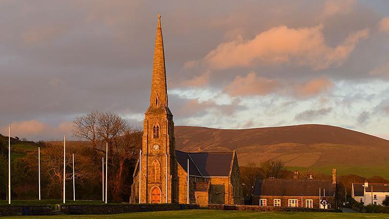 The exterior of the Royal Chapel, a church with a thin, tall steeple, the afternoon sun makes it glow, and flag poles are in front, with hills in the background.