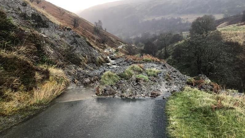 Rural minor road blocked by large mound of mud and stones which have slipped down from bank to the left