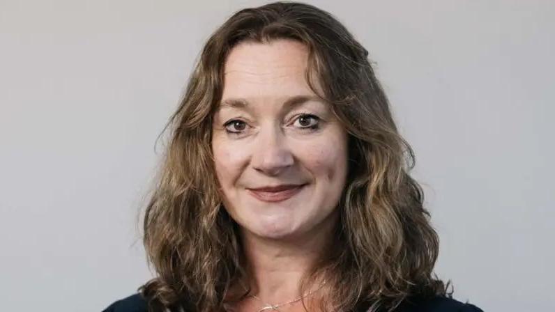 Daphne Caine, a woman with brown wavy hair smiles in front of a white backdrop.