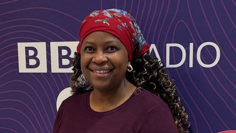 Rose Gicovi, wearing a red hair covering and a purple top in front of a purple BBC Radio CWR sign