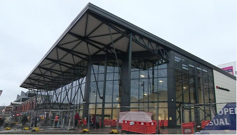 A large large glass building with a big canopy with barriers and railings in front.