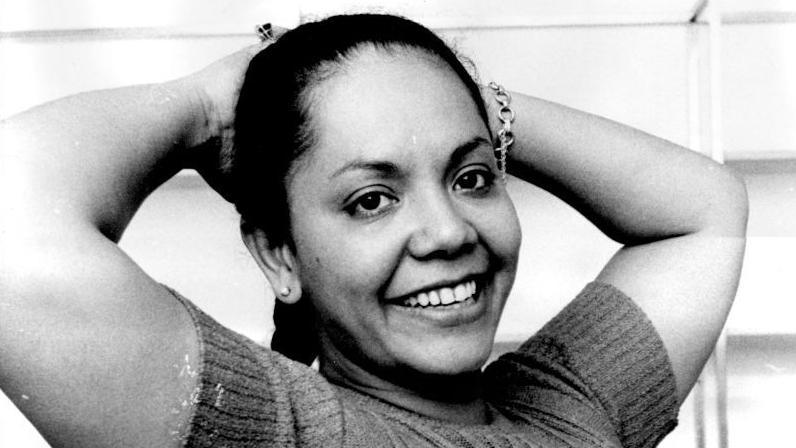 A black and white image of Candy Devine smiling with her arms behind her head as she wears a high-neck top.