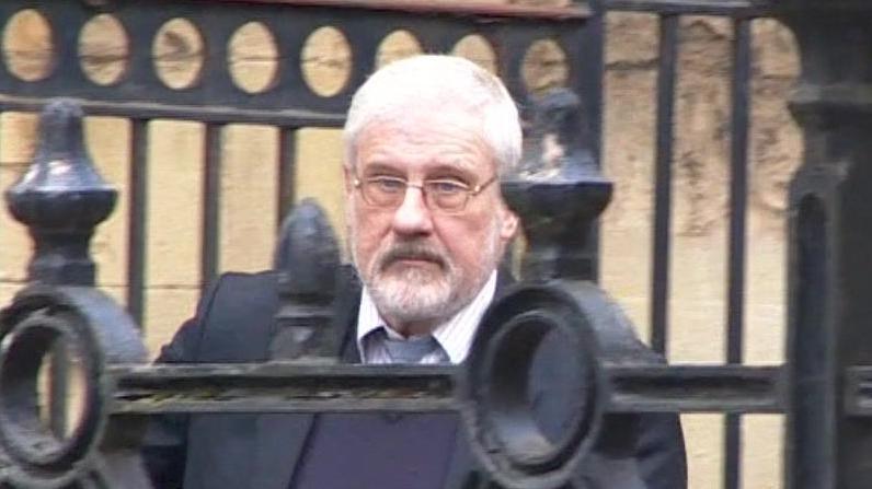 Stephen Cox, wearing a buttoned-up shirt and tie, walking out of Reading Crown Court, with the bars of railings in front of him as he walks down a ramp outside the building. He is wearing glasses and has short, white hair and a white beard.