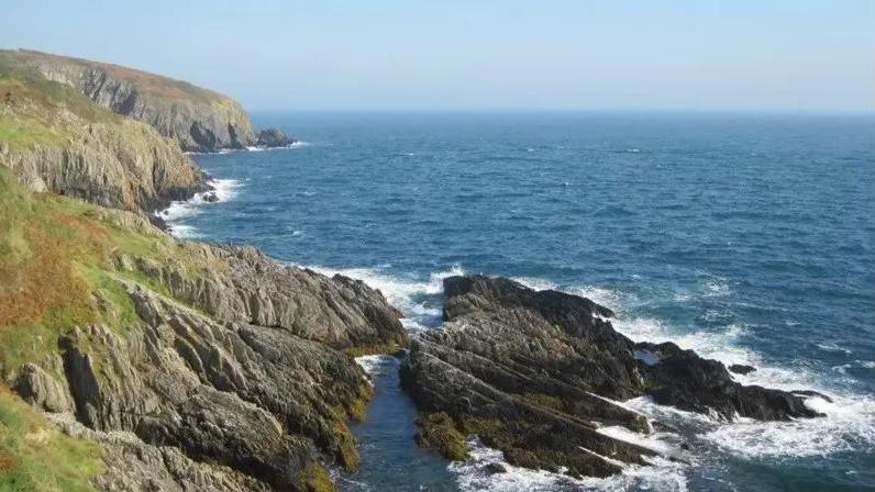 A coastline of rocky cliffs with green grass on top of them, with a large rocky outcrop below them and blue sea stretching off to meet the horizon.