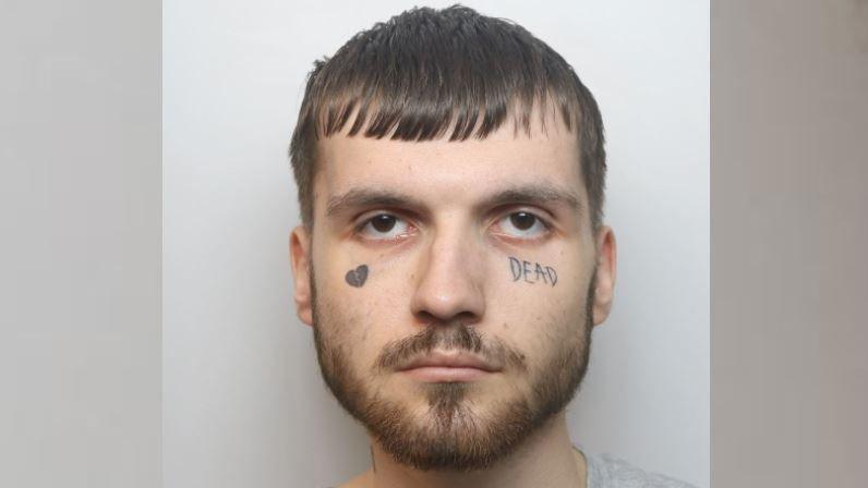 Custody image of Jack Towell. He has brown hair, brown facial hair and two facial tattoos. One is a broken love heart and the other is the word 'DEAD' under this left eye.