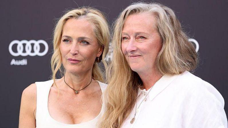 Gillian Anderson and Raynor Winn. Both are blonde, middle-aged women, both wearing white tops and standing next to each other on a red carpet with a black background smiling.