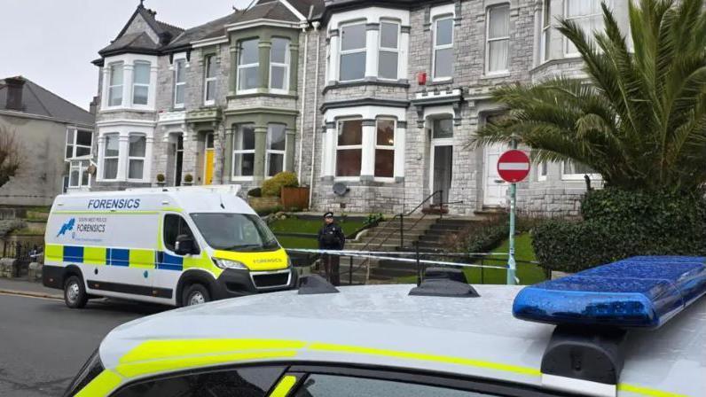 A police officer in uniform stands guard at a cordon outside a row of grey-bricked terraced houses in Plymouth. He is standing behind a line of police tape. A police forensics van is parked next to him on the road. The roof of a police car can also be seen.