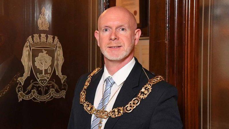 A bald man wearing a large gold chain, black suit jacket, white shirt and blue striped tie stands next to a wooden door which has the gold-embossed Glasgow coat of arms on it