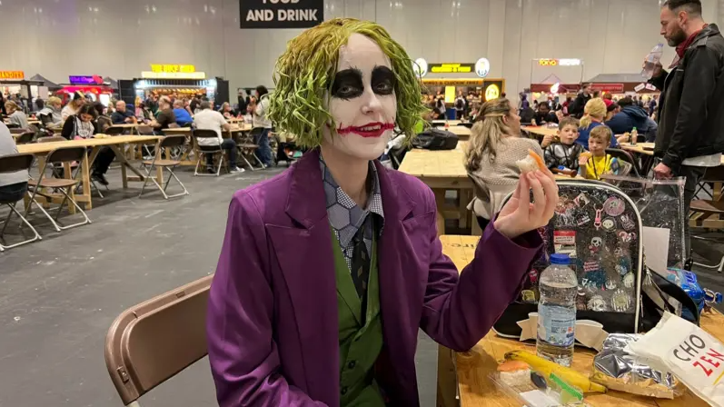 A young woman sat at a table eating sushi, dressed as the Joker with scraggly green hair and clown makeup, dressed in a purple suit with a green waistcoat.