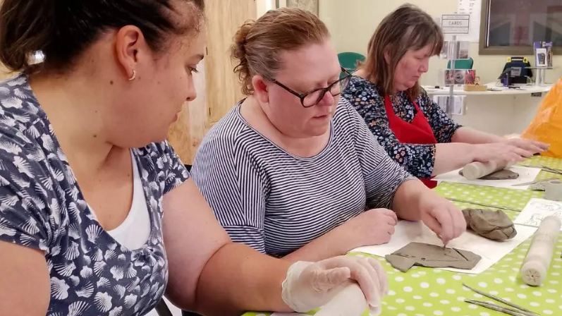 Three women rolling out clay or etching on to clay laid out flat on a table 