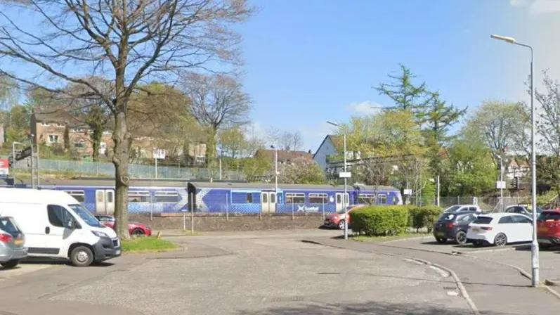 A general view of the car park next to the crescent in Dalmuir. A train going on tracks behind a fence is in the centre of the image. Cars are parked on both sides of a short road with a tree on the left side. It is a sunny day.