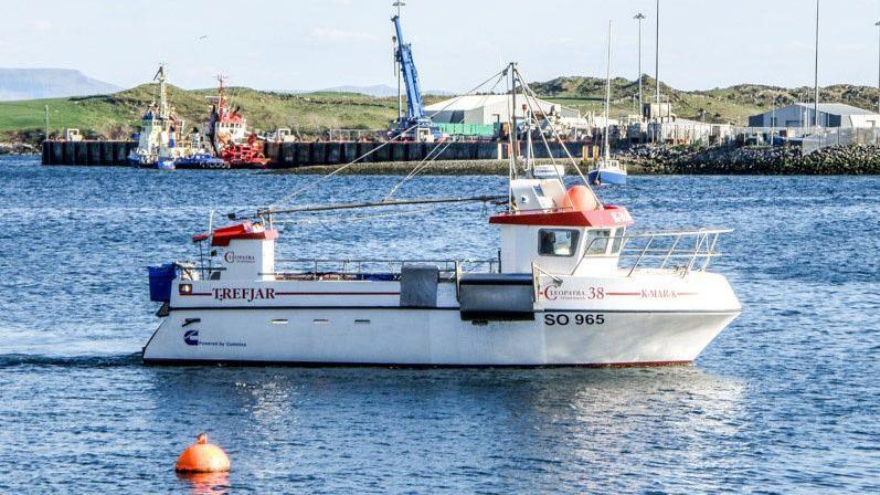 Kevin McCloskey's fishing boat, the FV K-Mar-K, on the water near a rural harbour on a sunny day. The boat is white with red branding. There is a red canopy over the wheelhouse and there is a metal safety rail around the hull.