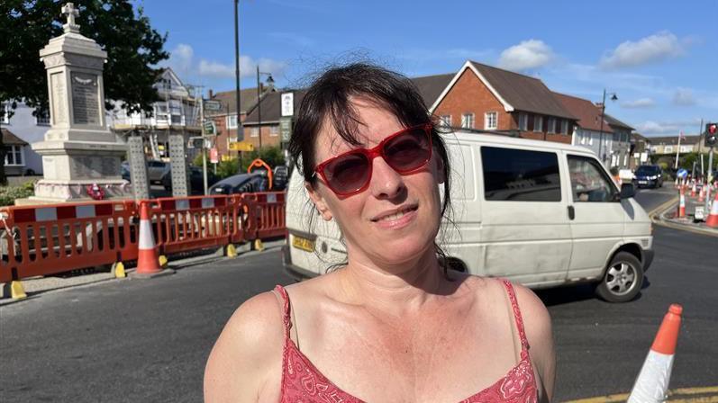 Councillor Ellen Neville stands on the corner of Hawkhurst crossroads with a white van passing behind her. She has dark-hair loosely tied back, and is wearing red-rimmed sunglasses and a red top.