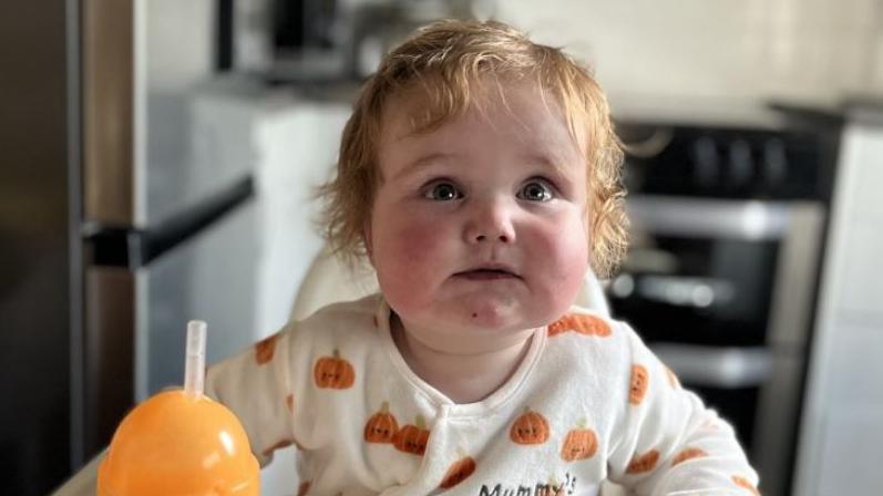 A toddler in his high chair. He has wispy blond hair and wears a cream babygro with orange pumpkins on it. An orange sippy cup is next to him and an oven can be seen behind.