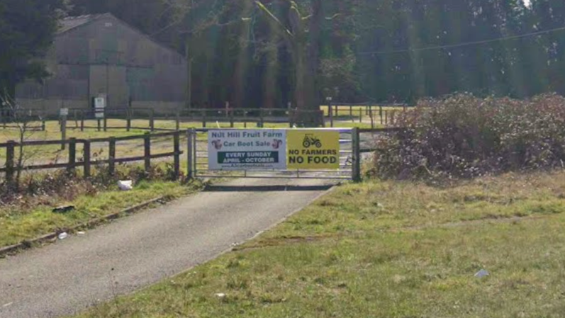 The exterior of a farm next to the A3 near Guildford. On a fence there are two signs. One says 'Nut Hill Fruit Farm Car Boot Sale. EVERY SUNDAY APRIL - OCTOBER.' The other says 'NO FARMERS, NO FOOD.'