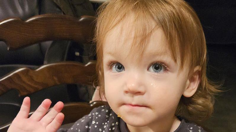 A two-year-old girl with shoulder-length brown hair and blue eyes is wearing a dark top with white stars on it. She is sitting on a chair and is holding up her right hand