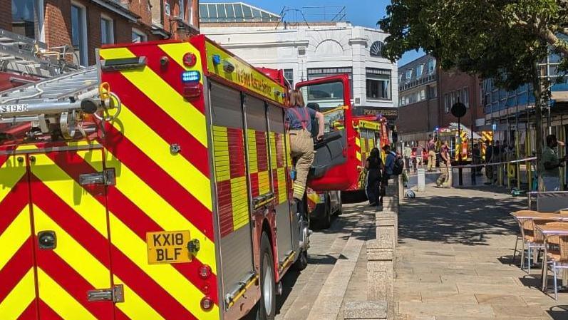 View behind fire engines stood in the middle of a road which is on the left. A woman is climbing into the fire engine. In the distance, firemen are standing in the middle of a road which is cordoned off with tape.