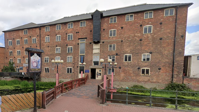 Haven Mill - a four-storey red-brick building. There is a pub sign outside reading Tap 'n' Spile and at the front of the building is a brick bridge with red fencing either side over a pond