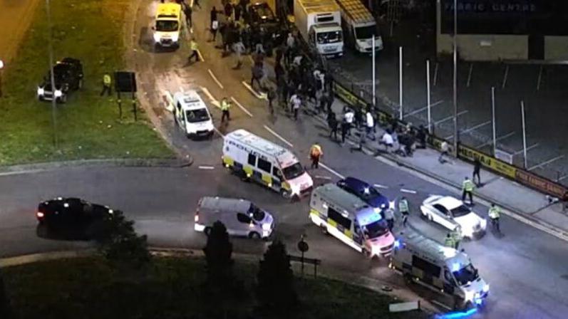 An aerial view of several police cars and vans driving down a road, with a crowd of people walking along the nearby pavement.