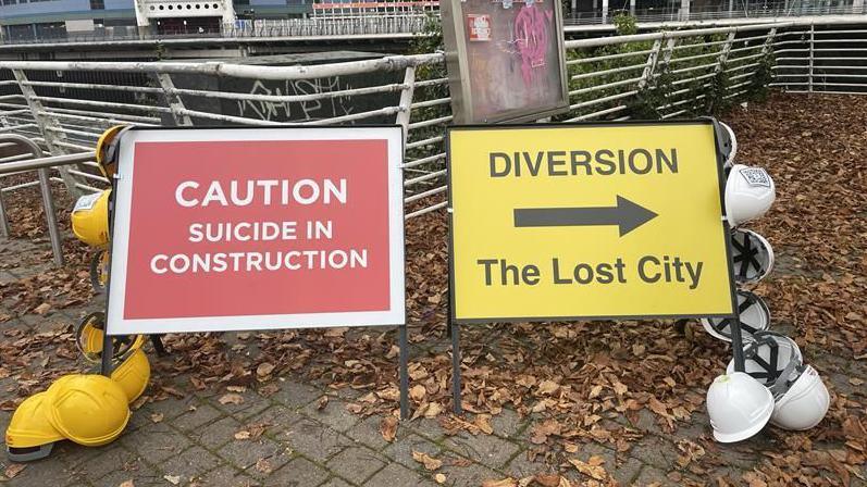 Official-looking road signs, one red and one yellow. The red one says: CAUTION Suicide in construction, and the yellow one says DIVERSION ---> The Lost City. The signs have hard hats on either side of them, with a small QR code stuck to the top of each side. 