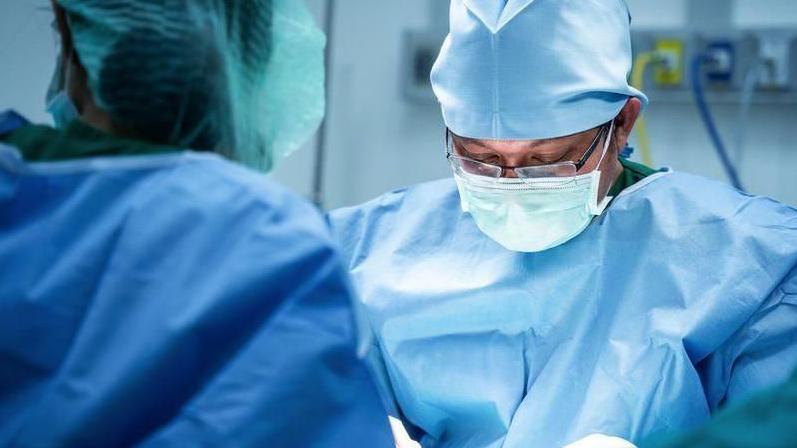 Close up of a surgeon during an operation. He is wearing blue scrubs and a face mask. He is leaning slightly forward and looking down through a pair of black-framed spectacles. 