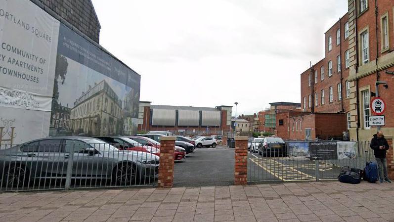 A small, open, ground-level car park with a three-storey red brick hotel on one side and a large property advertisement on the other. A metal fence runs across the entrance-side, bordered by two brick columns.