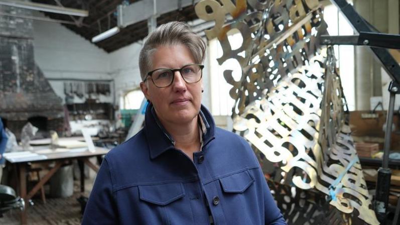Kate Holvey-Williams is wearing glasses and has short hair styled into a slight quiff. she is standing against the letters memorial in the forge.