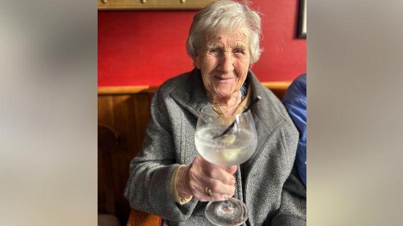 Image of Hilda in a grey coat holding up a gin and tonic