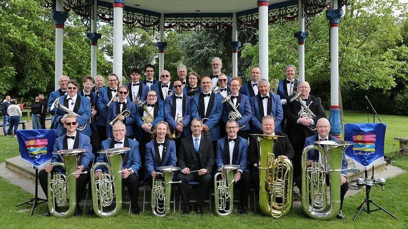 A brass band lined up in four rows in front of a bandstand. Members are holding their instruments and wearing matching uniforms of blue blazers with bow ties.